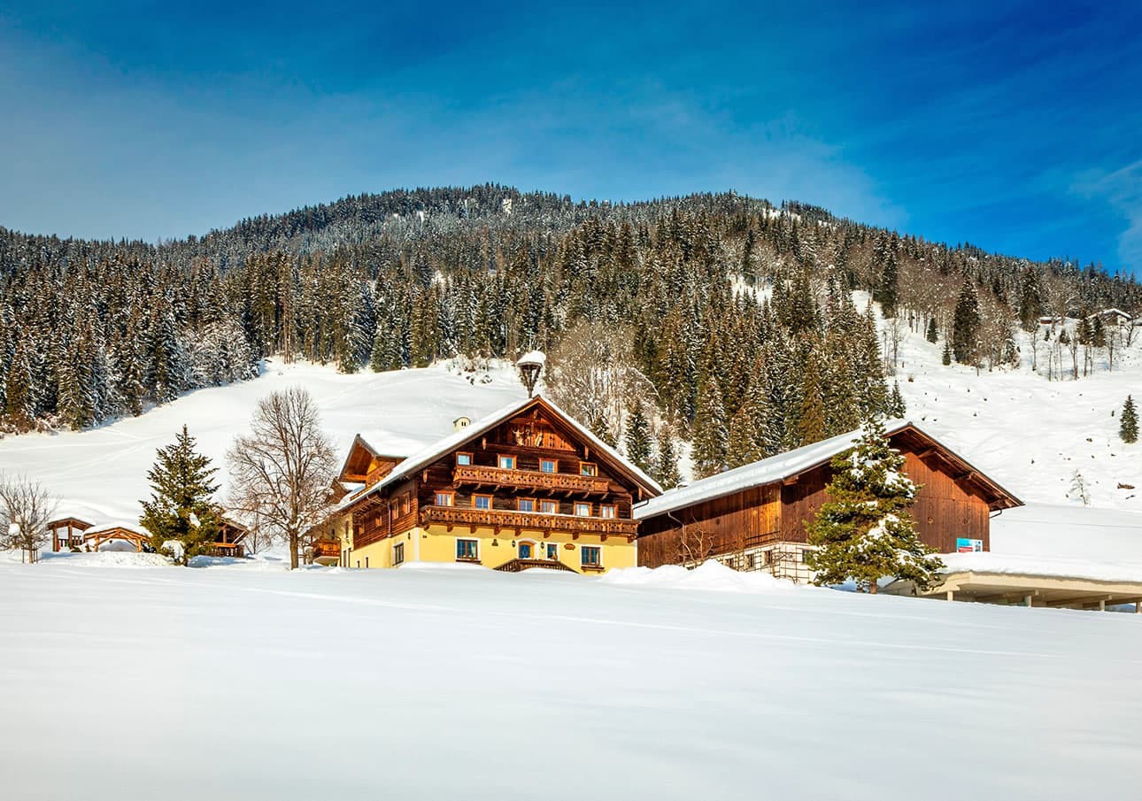 Snow-covered wooden houses in front of a wooded hill under a blue sky.