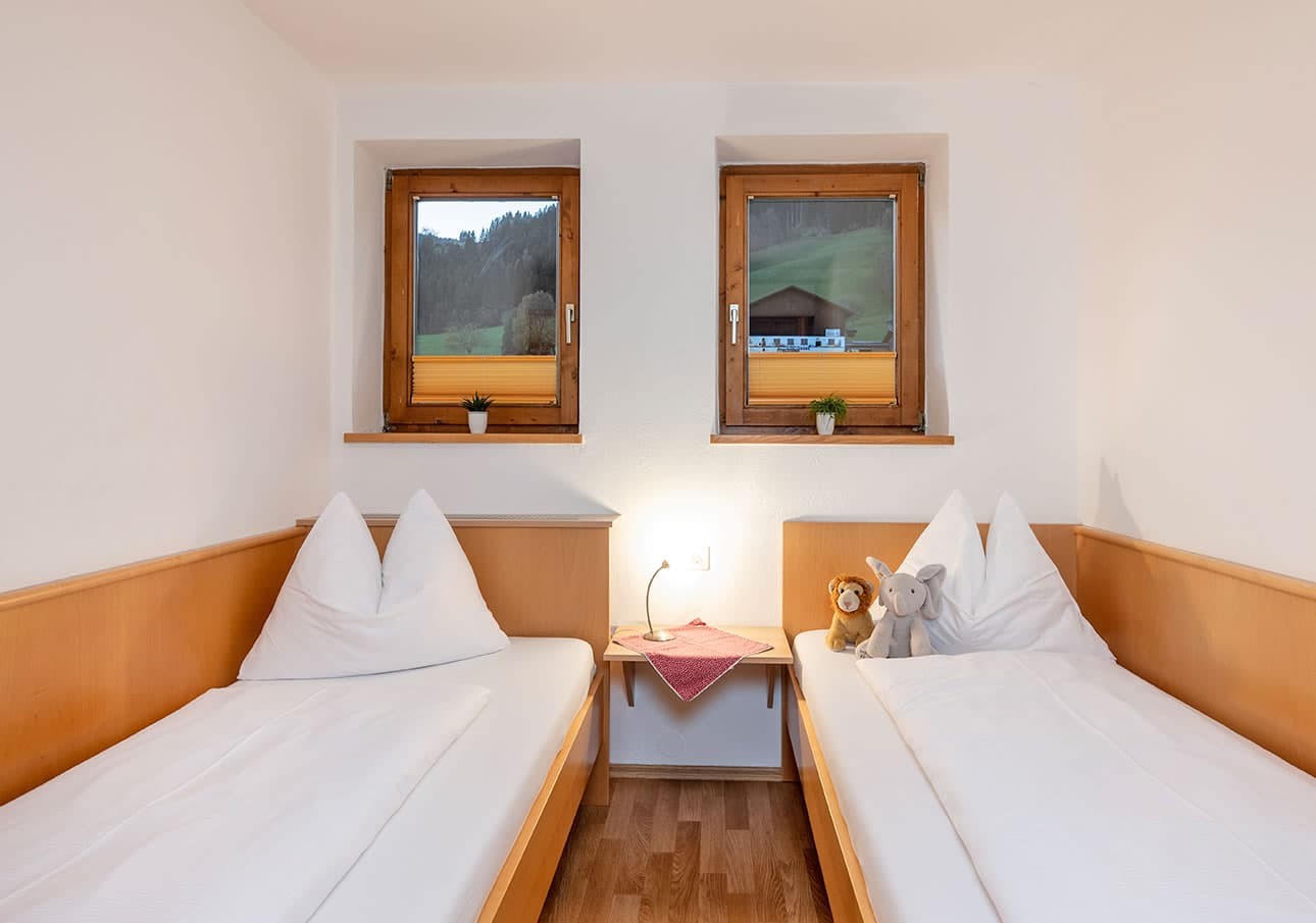 Two neatly made single beds with plush toys, under two windows in a cozy room