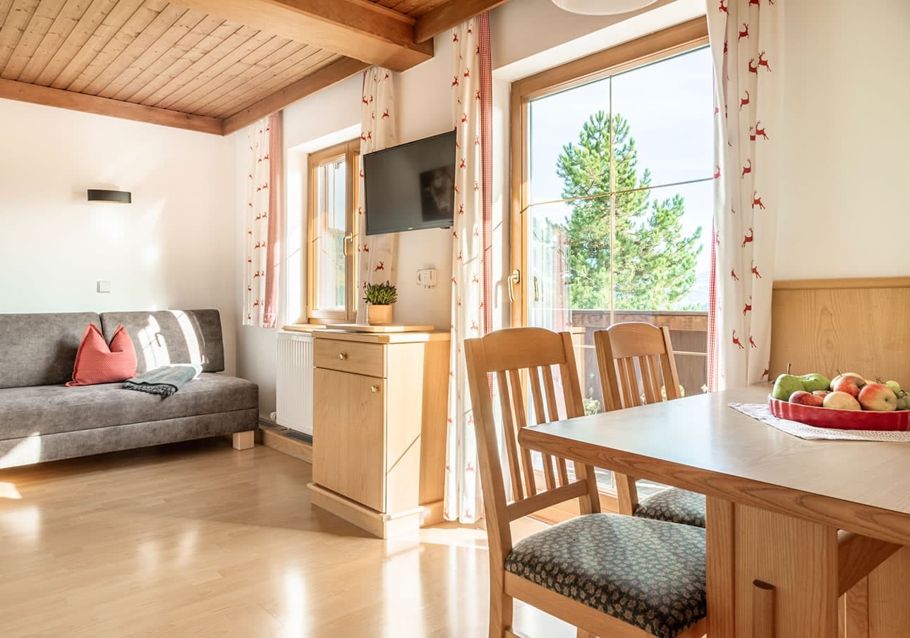A cozy living room with wooden furniture, a sofa, and large windows showing greenery outside