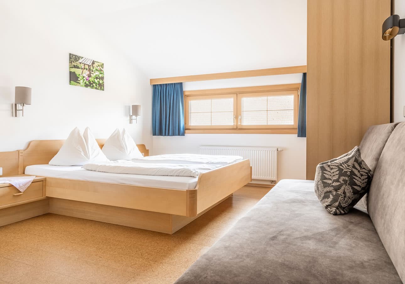 Bright, minimal bedroom with a wooden bed, blue curtains, and a gray sofa
