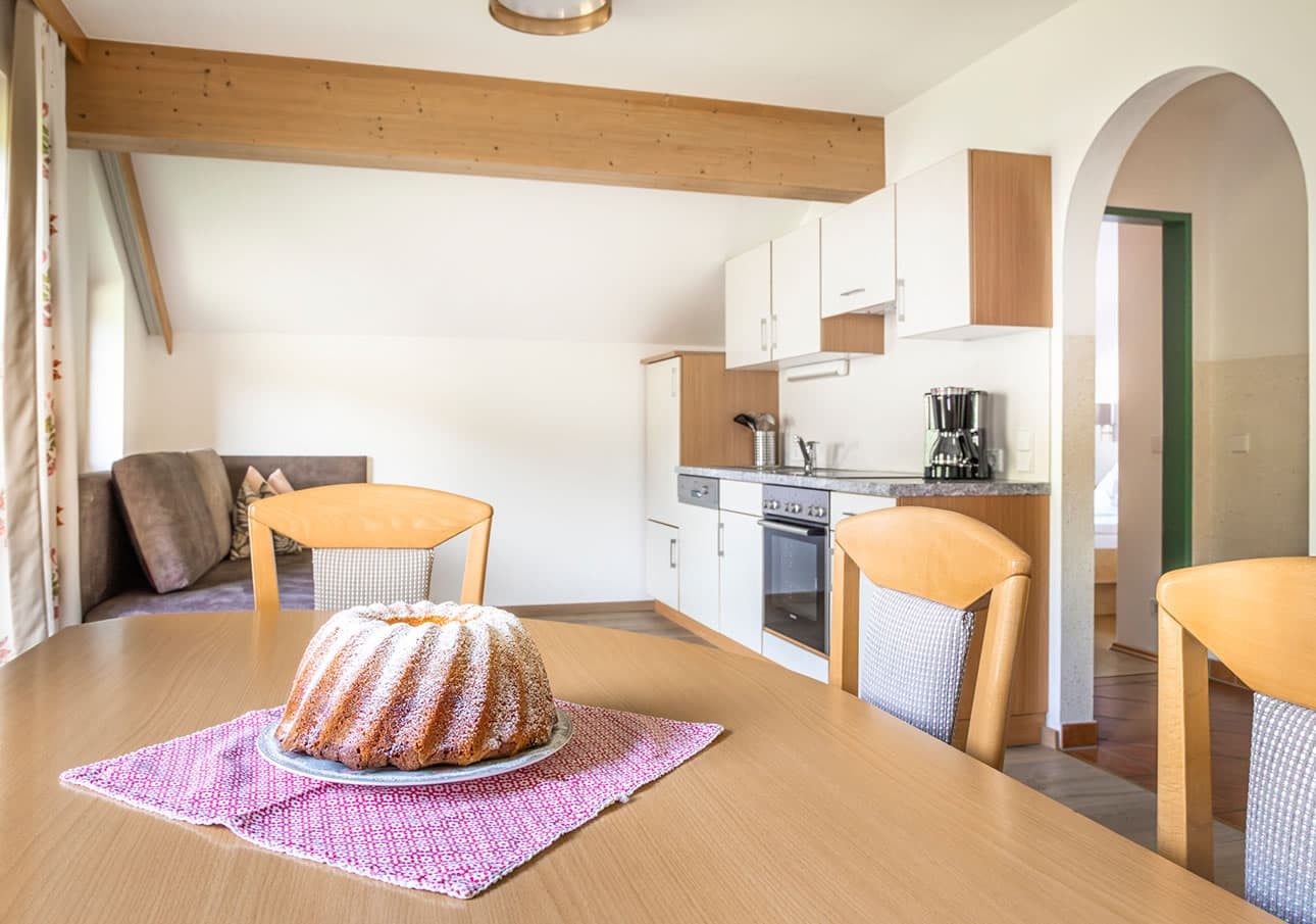 A cake sits on a table in a cozy kitchen with wooden chairs and white cabinets