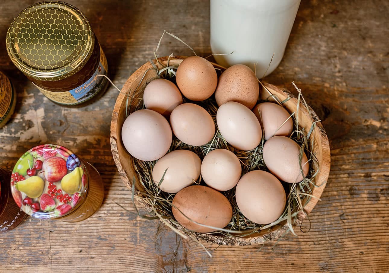 A wooden bowl of eggs on hay next to jars and a bottle on a wooden surface