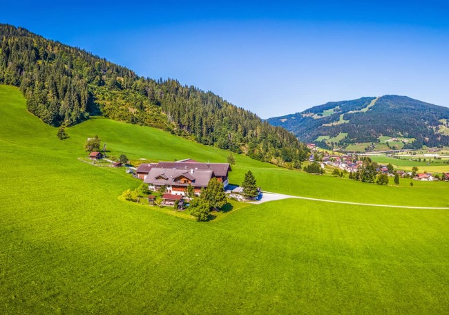 Landschaft mit Prechtlhof auf grüner Wiese vor bewaldeten Hügeln und blauem Himmel