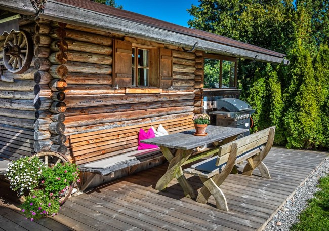 Holzhütte mit Veranda, Holzbank, Tisch und Grill, umgeben von Pflanzen