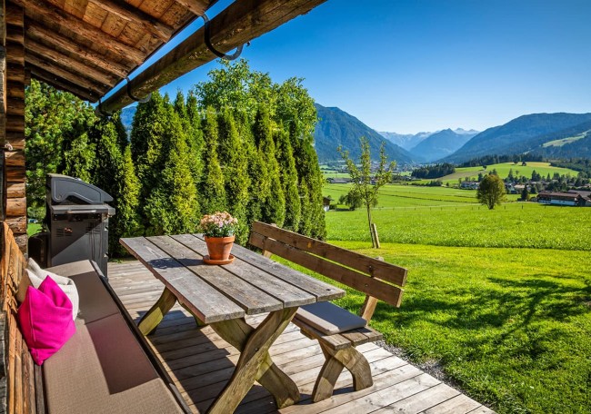 Holzterrasse mit Tisch und Sofa, umgeben von grünen Wiesen und Bergen unter blauem Himmel