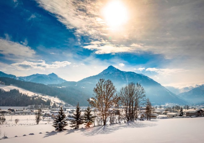 Verschneite Landschaft mit Bäumen und Bergen unter strahlender Sonne und blauem Himmel