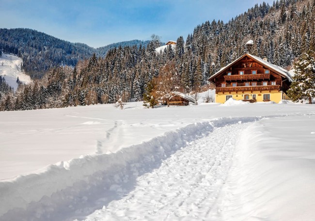Ein schneebedeckter Pfad führt zu einem traditionellen Prechtlhof vor einer bewaldeten Berglandschaft