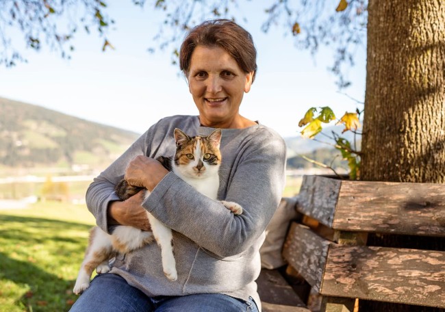 Annemarie Rettensteiner hält eine Katze auf einer Bank im Freien, lächelnd in die Kamera