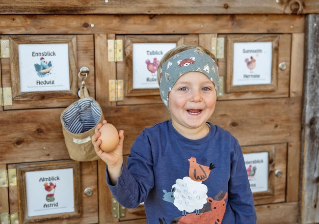 Ein lächelndes Kind hält ein Ei vor einer Holzwand mit Hühnerbildern