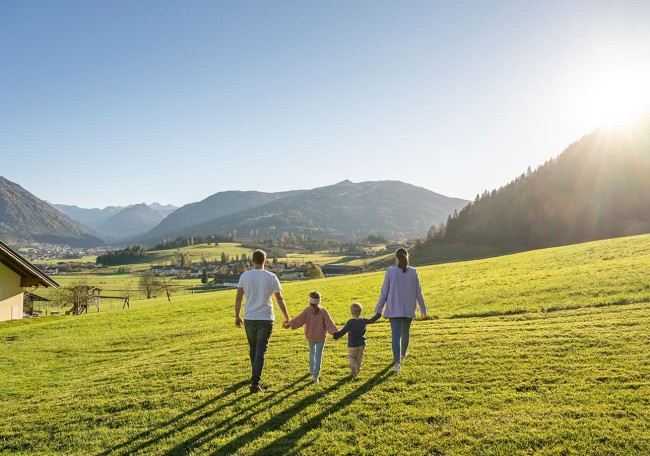 Familie spaziert über grüne Wiese mit Bergen im Hintergrund bei Sonnenuntergang