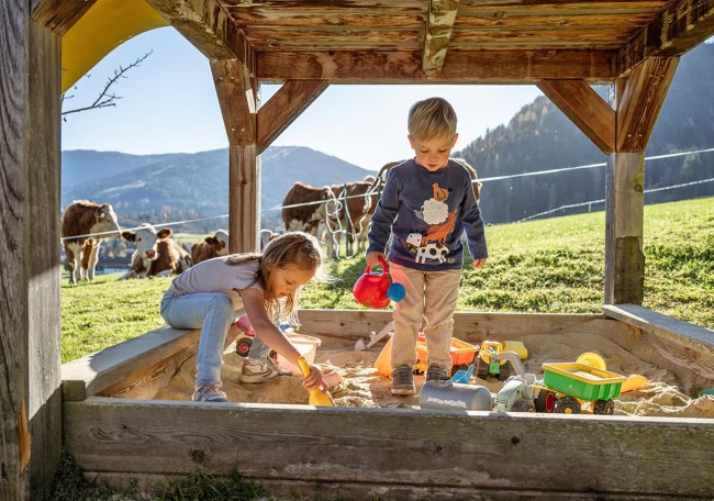 Kinder spielen im Sandkasten, umgeben von Wiesen und Kühen, bei sonnigem Wetter