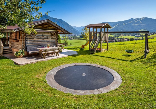 Holzhaus mit Spielplatz und Trampolin in einer grünen Berglandschaft bei sonnigem Wetter