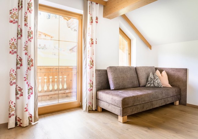 Wohnraum mit braunem Sofa, Holzboden und großen Fenstern mit floralen Vorhängen