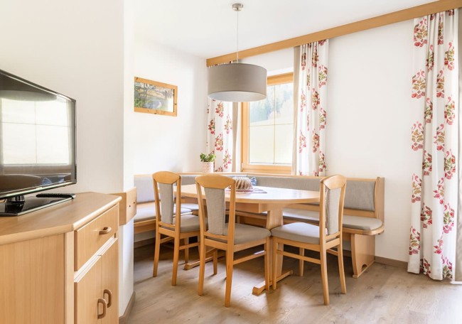 Helle Essecke mit Holzstuhlgruppe und Eckbank, neben einem Fenster mit floralen Vorhängen