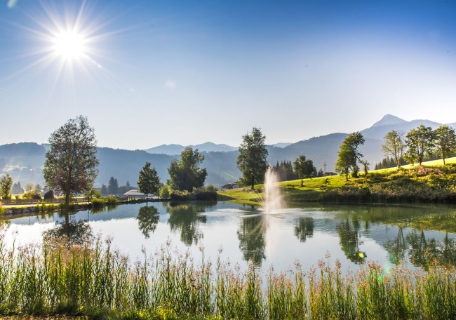 Ein klarer See in Flachau mit Bäumen, Bergen und einer Wasserfontäne im Sonnenlicht