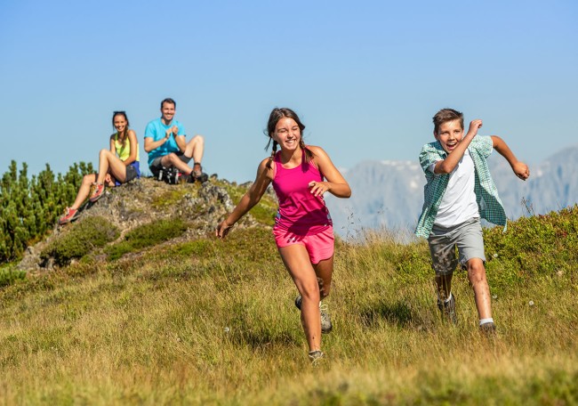 Kinder laufen auf einer Wiese, während zwei Erwachsene im Hintergrund sitzen