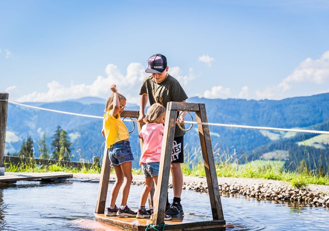 Drei Kinder balancieren auf einem Holzbrett über einem flachen Wasserbecken in der Natur