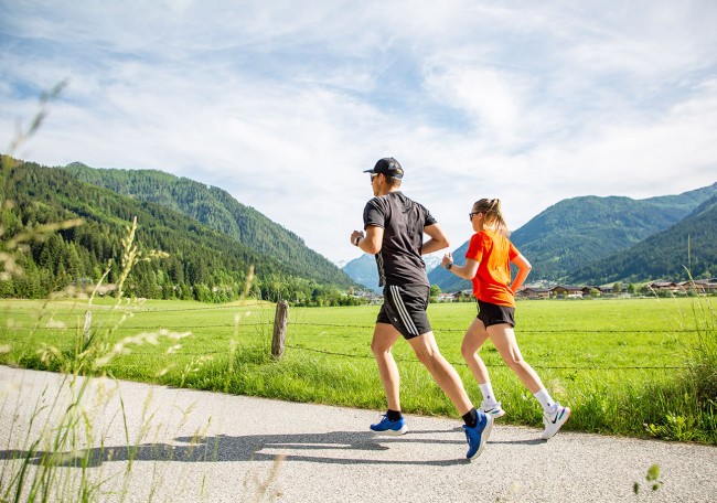 Zwei Menschen joggen auf einem Weg in einer grünen, bergigen Landschaft unter blauem Himmel