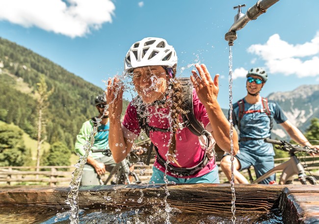 Frau erfrischt sich an einem Brunnen während einer Fahrradtour in bergiger Landschaft