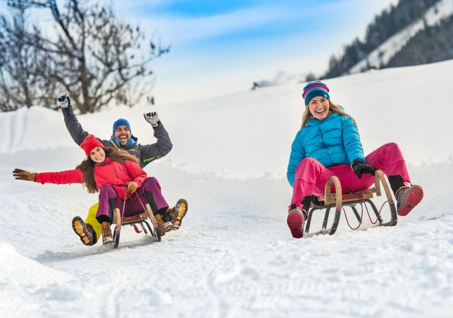 Personen fahren lachend auf Schlitten durch eine verschneite Winterlandschaft