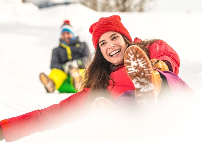 Zwei Personen rodeln fröhlich im Schnee und tragen bunte Winterkleidung