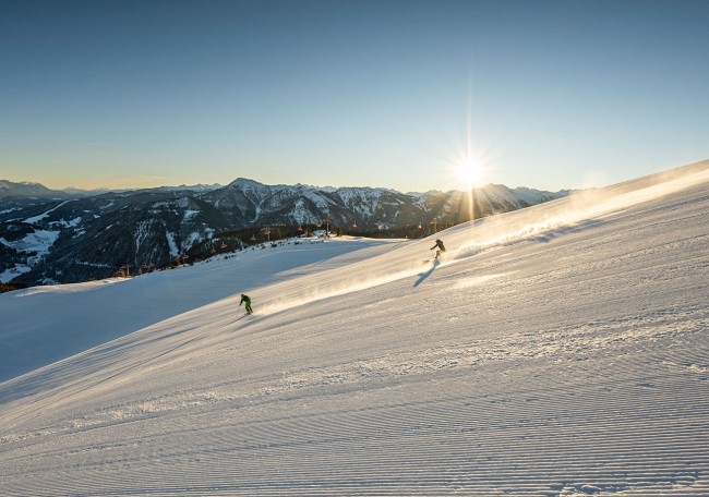 Zwei Skifahrer fahren bei Sonnenaufgang eine schneebedeckte Piste hinunter