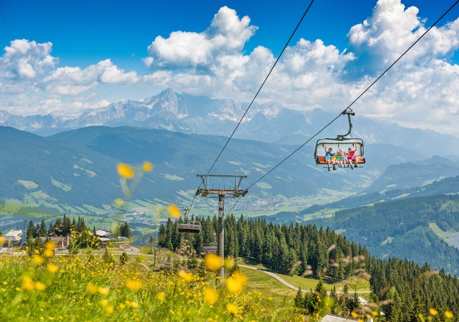 Eine Seilbahn überquert grüne Wiesen mit Bergen im Hintergrund