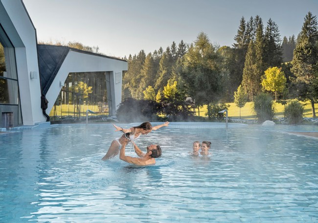 Eine Familie spielt fröhlich gemeinsam in einem dampfenden Außenpool vor einer Waldkulisse