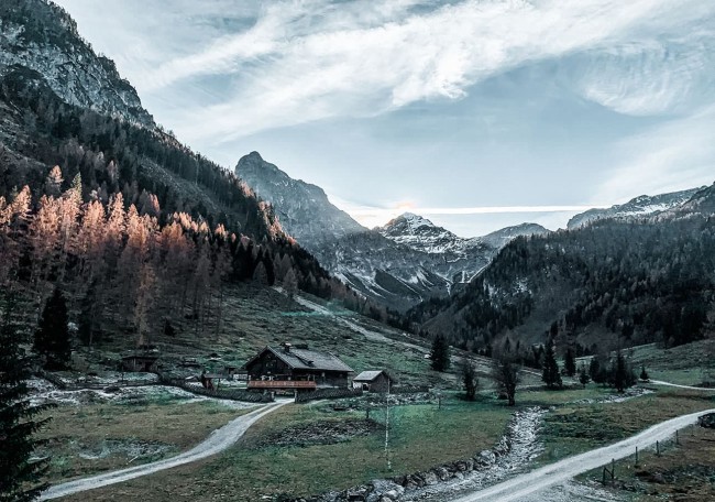 Berglandschaft mit verschneiten Gipfeln, Wäldern und einer Hütte im Tal bei Sonnenaufgang