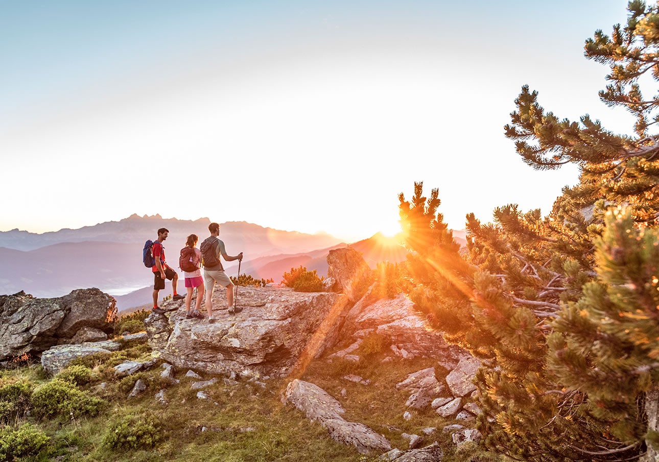 Three hikers stand on rocks, admiring a sunset over mountains and trees